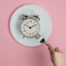 Breakfast time! Female hand with fork about to eat from a plate with a retro alarm clock on pink pastel background. Top view